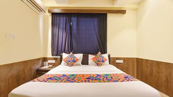 Habitación de hotel con cama, mesita de noche, toma de corriente y ventana con cortinas.