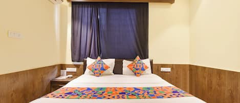 A hotel room with a bed, a nightstand, a power outlet, and a window with curtains.