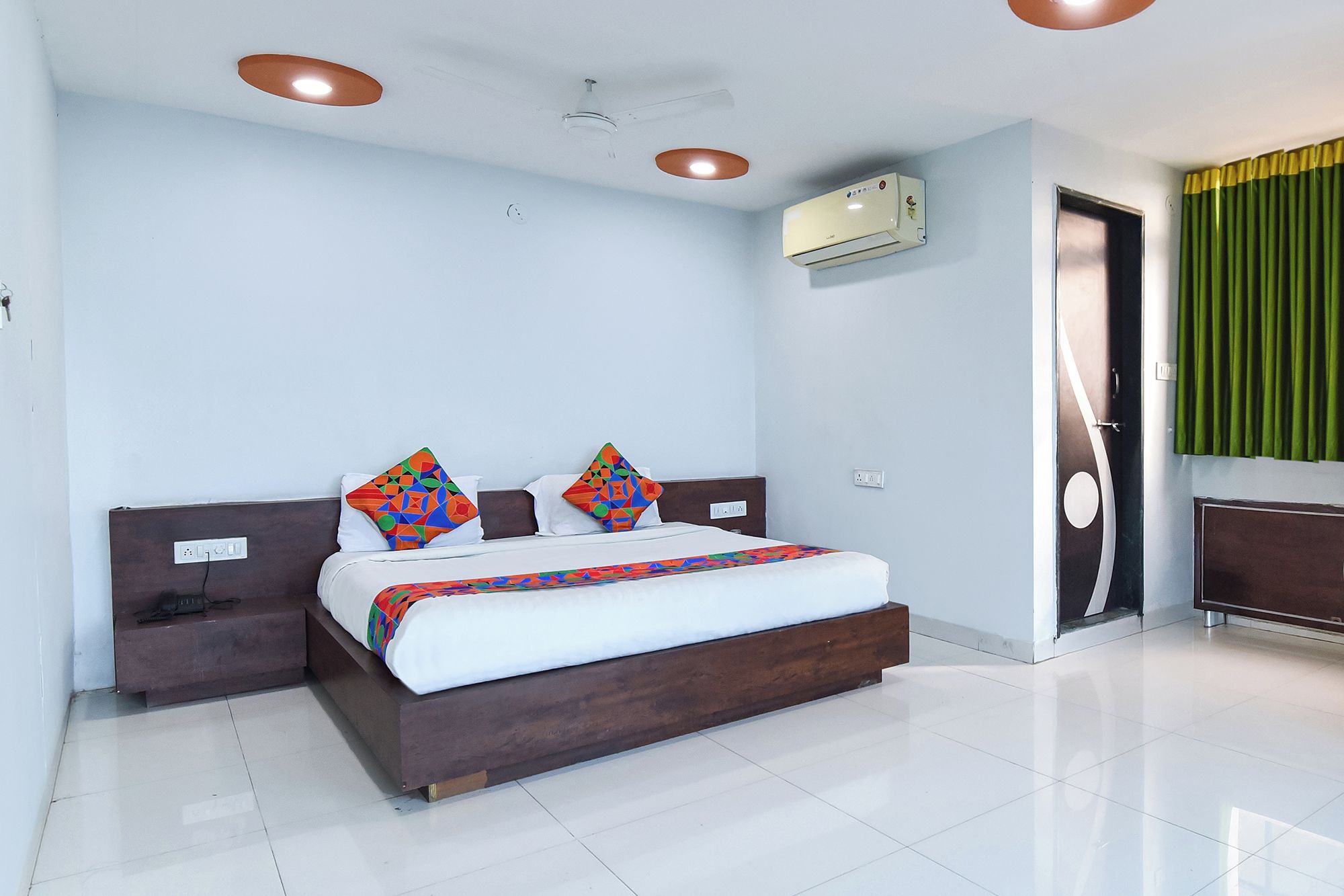 A modern hotel room with a large bed, two bedside tables, a wall-mounted air conditioner, and a green curtain.