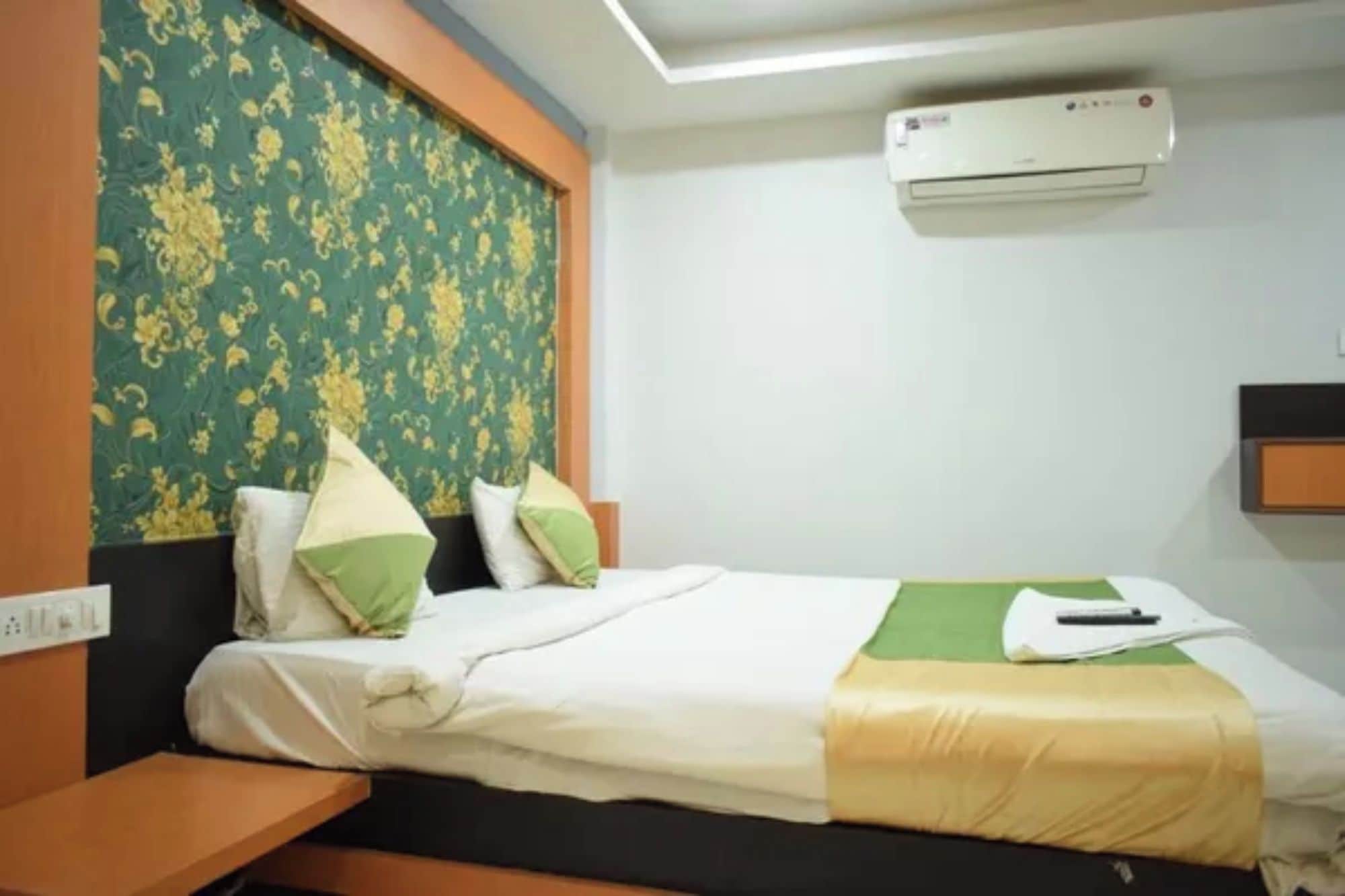 A hotel room with a bed, a nightstand, a wall-mounted air conditioner, and a patterned wallpaper.