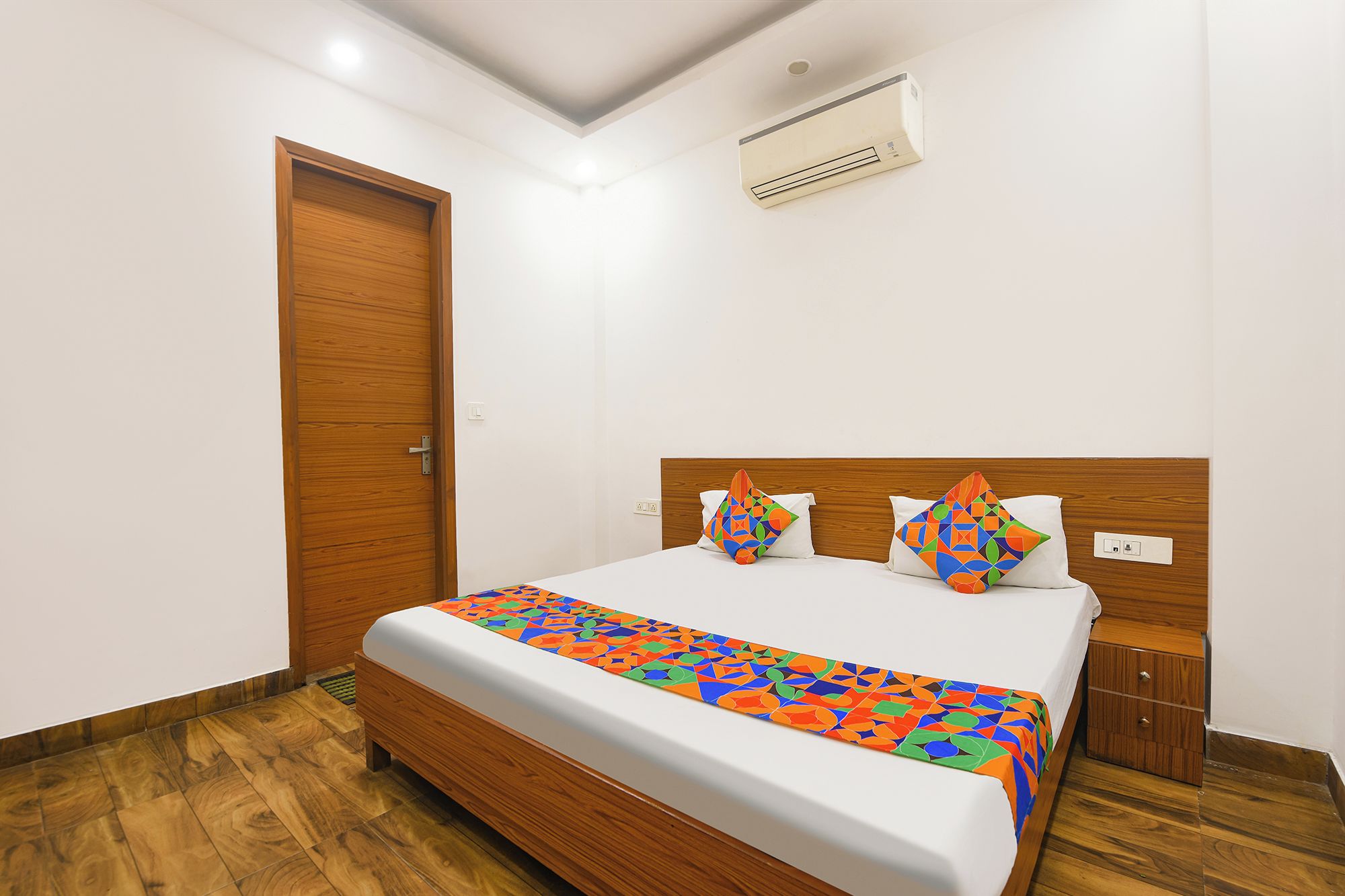 Ein Hotelzimmer mit einem hölzernen Bett, einem weißen Kopfteil und einer farbenfrohen Bettdecke mit geometrischem Muster.