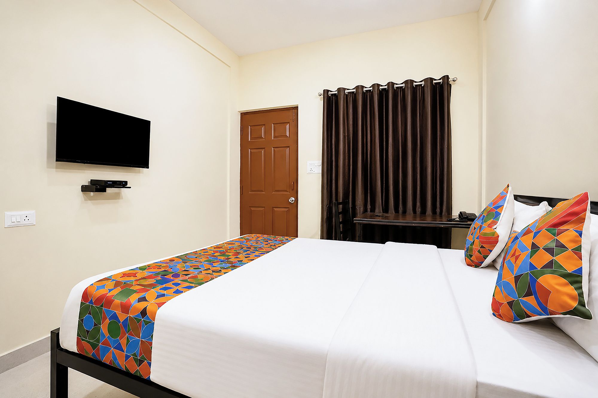 Una habitación de hotel con una cama, un televisor montado en la pared, una puerta y una cortina.