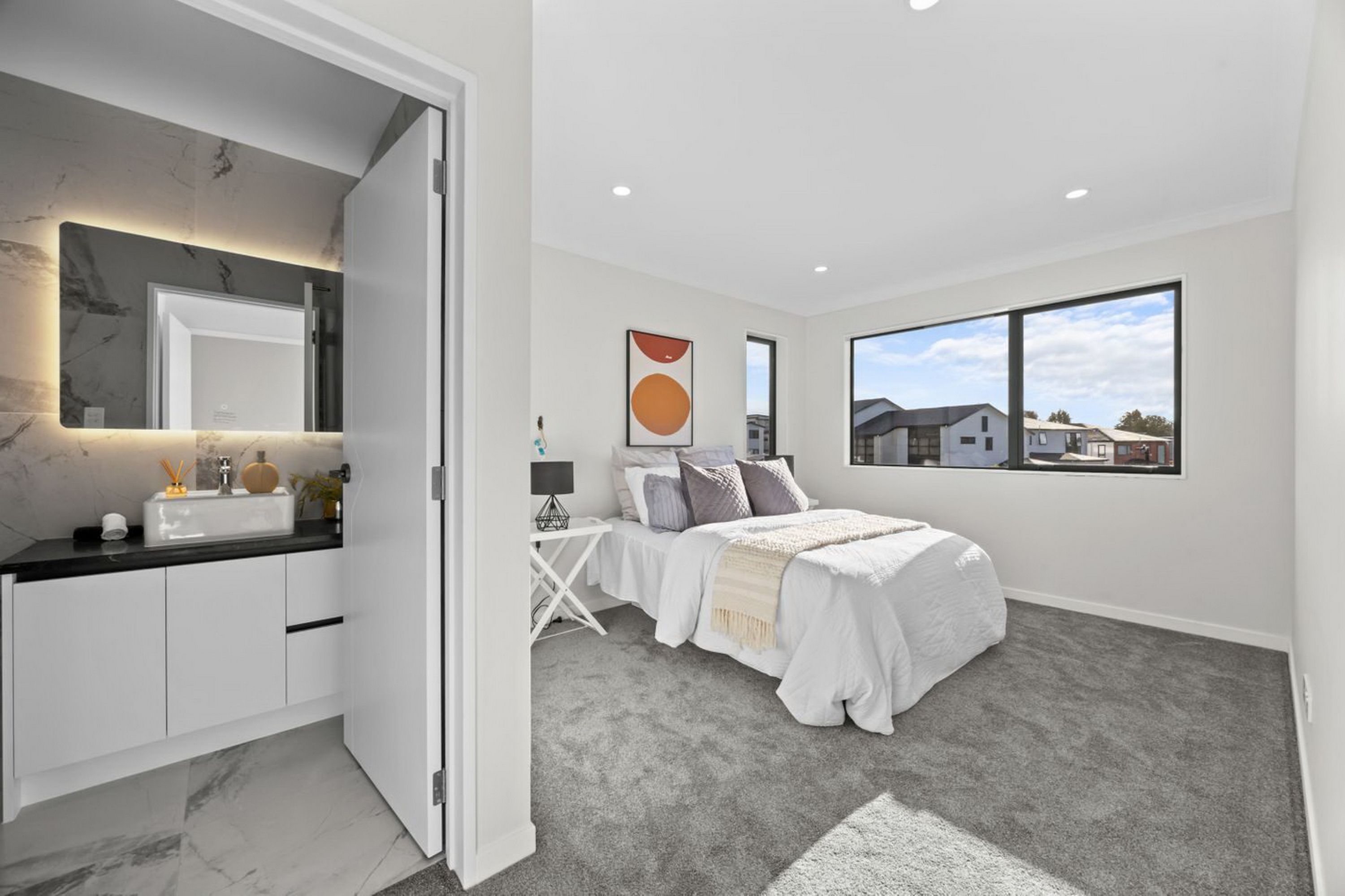 Un dormitorio moderno con una cama grande, una mesita de noche, un tocador, una ventana con vistas a casas y un baño con espejo y lavamanos.
