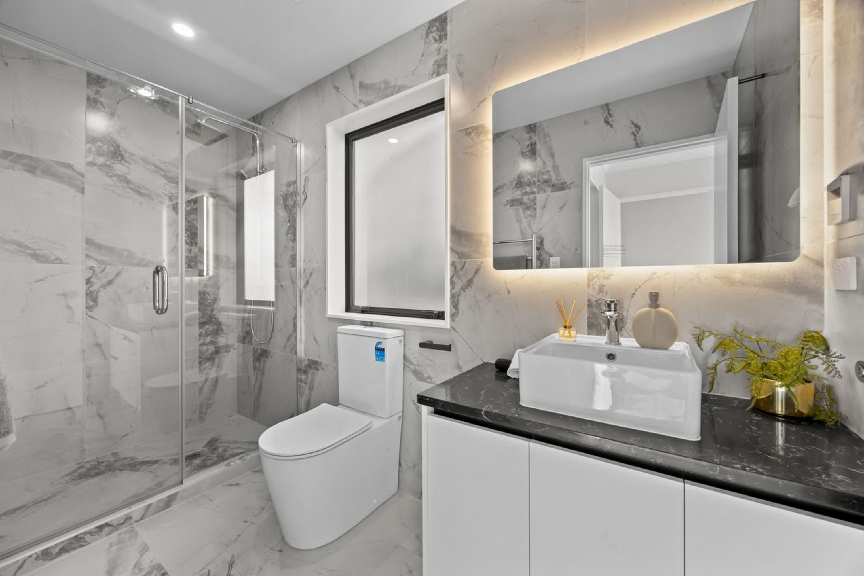 A modern bathroom with a glass shower enclosure, a white toilet, a sink with a marble countertop, and a large mirror.