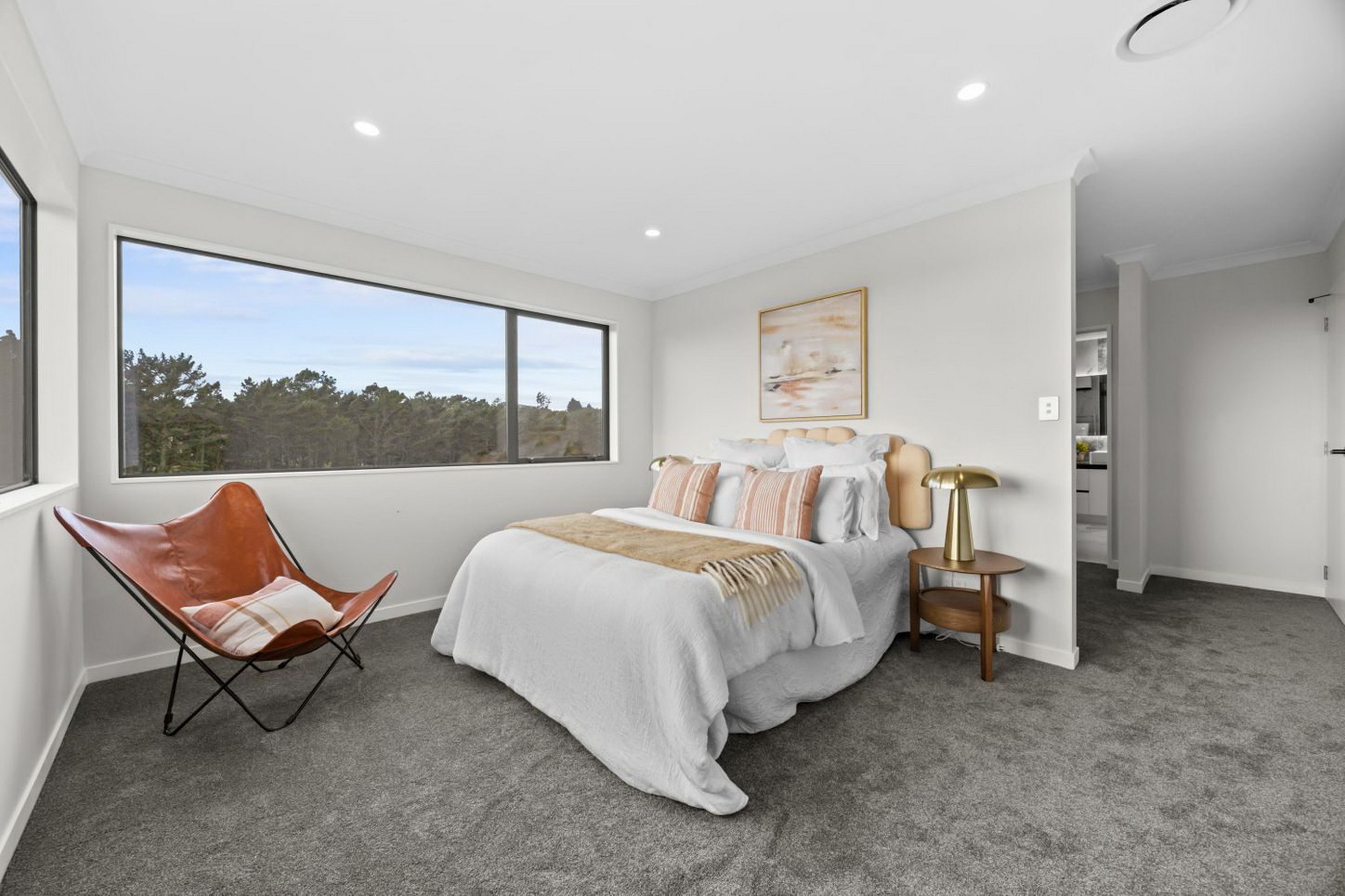 A bedroom with a large window, a bed with multiple pillows, a chair, and a bedside table with a lamp.