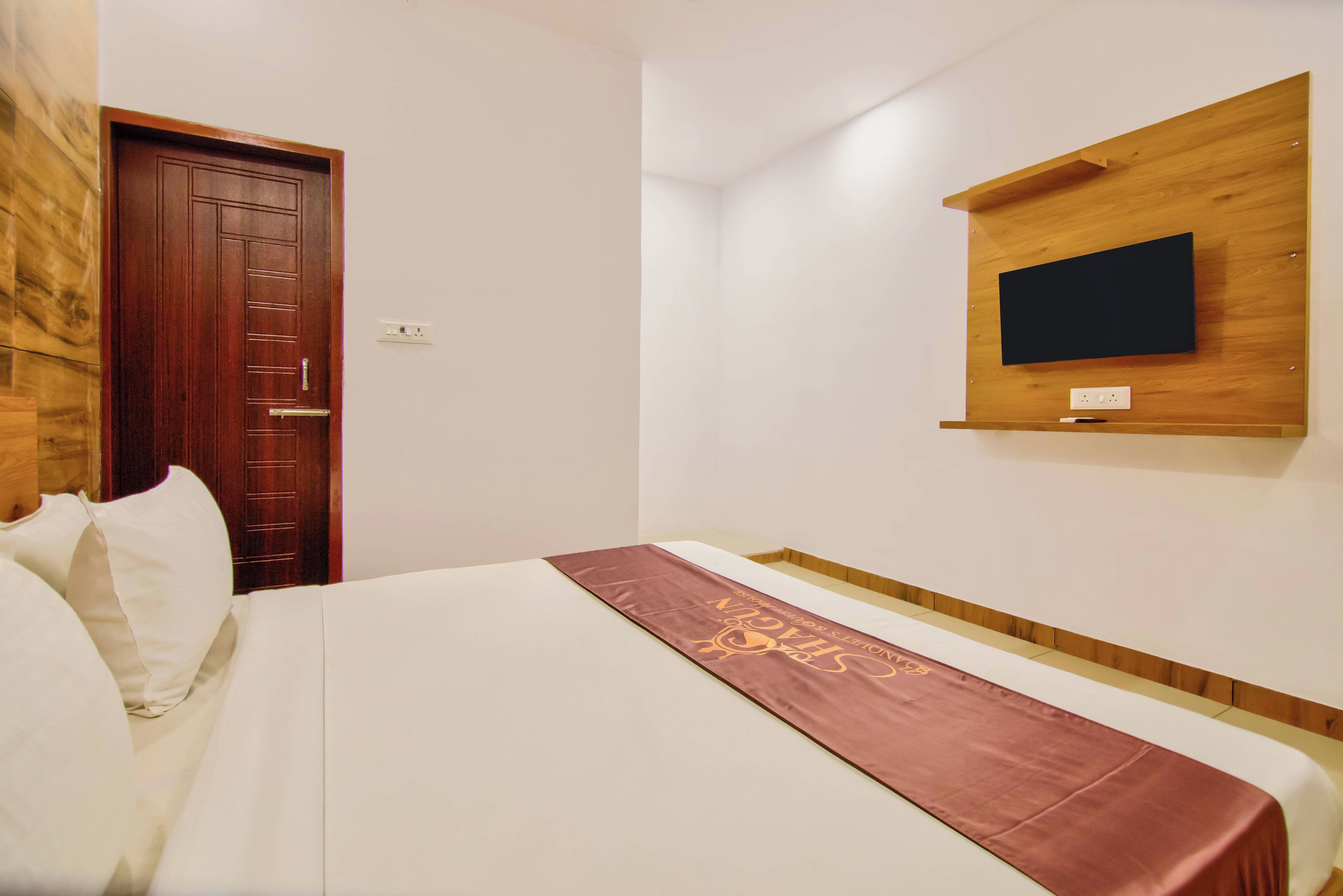 Una habitación de hotel con una cama, una puerta de madera, un televisor montado en la pared y un estante adosado.