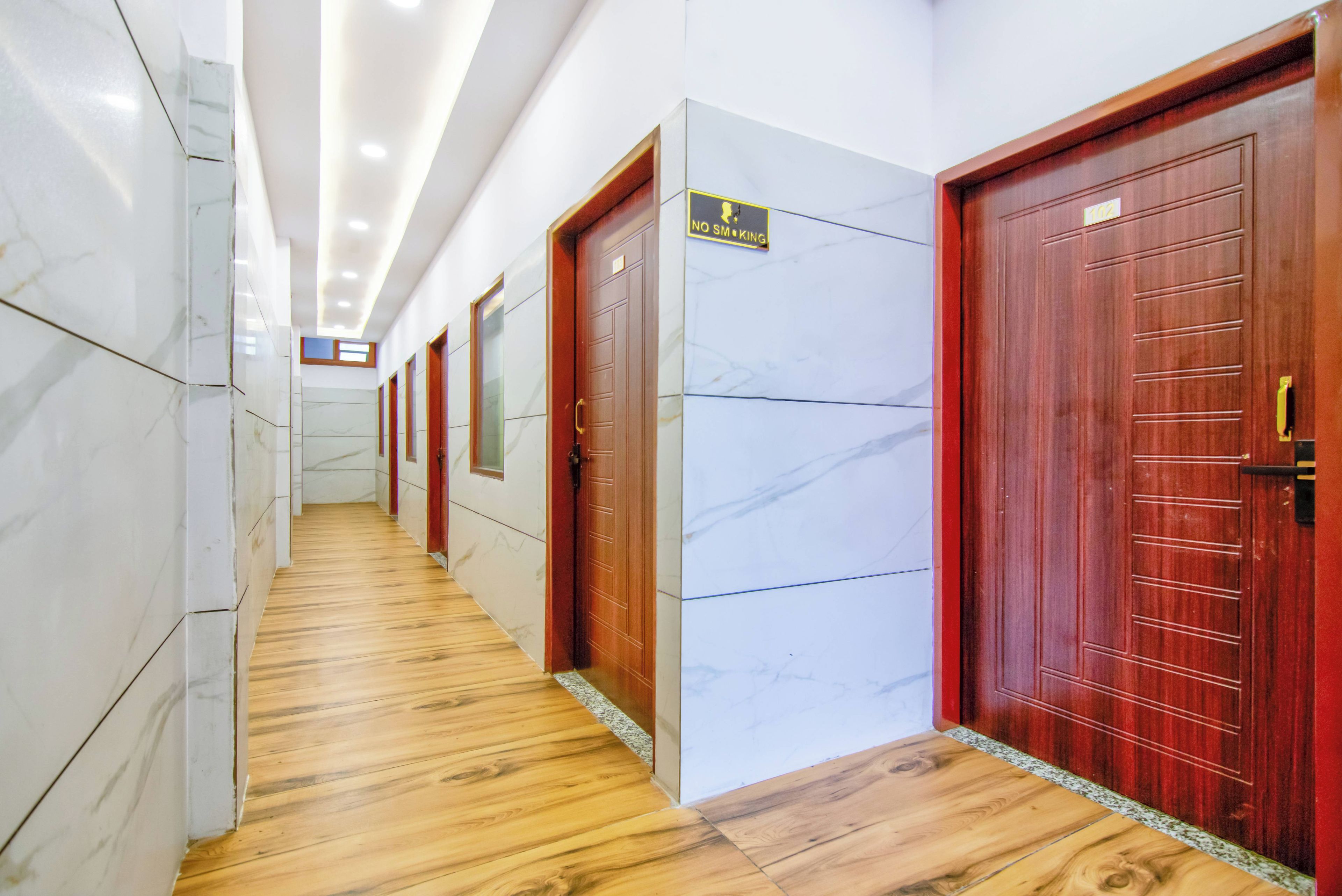 Pasillo de hotel con suelo de madera, paredes de mármol blanco y puertas de madera con un cartel que indica 'No Smoking'.