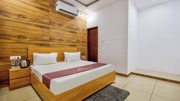 Habitación de hotel con cabecera de madera, cama con ropa blanca, mesita de noche con tetera y aire acondicionado de pared.