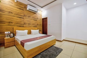 Habitación de hotel con cabecera de madera, cama con ropa blanca, mesita de noche con tetera y aire acondicionado de pared.
