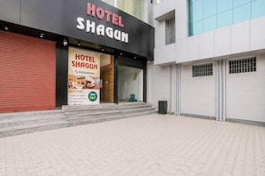 Entrada de un hotel con un letrero que dice 'HOTEL SHAGUN' y un anuncio de 'HOTEL SHAGUN DELUXE ROOMS' que muestra un número de teléfono y servicios como 'Amplias y elegantes', 'Opciones con y sin aire acondicionado' y 'Acceso a Wi-Fi gratuito'.