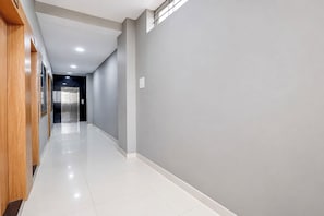 Un pasillo largo y estrecho con una puerta de madera a la izquierda y un ascensor de vidrio a la derecha.