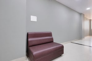 Un área de espera con un banco de cuero marrón sobre una pared gris, piso blanco y una lámpara en el techo.