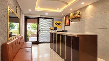 Área de recepção de hotel, com balcão de atendimento, espaço de estar com sofá, ventilador de teto e parede decorativa com quadros.