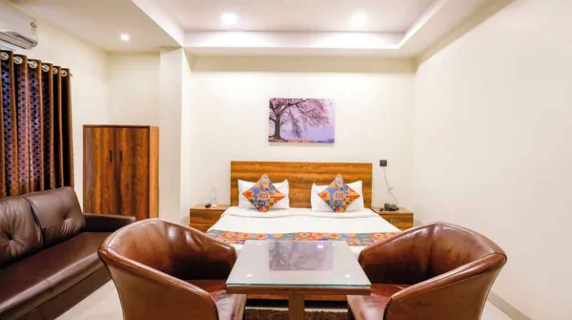 Habitación de hotel con cama, mesita de noche, armario de madera, sofá de cuero marrón y mesa de vidrio con dos sillas.
