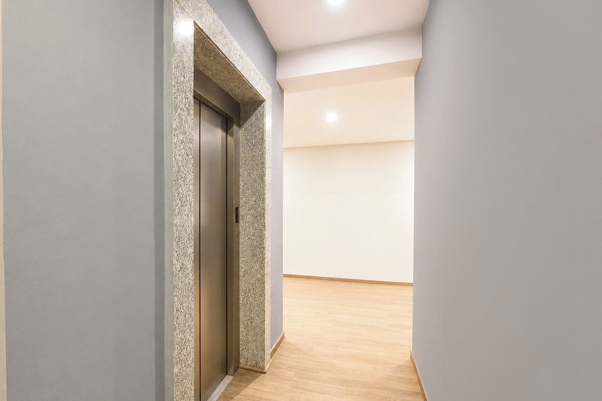 Passadís amb paret amb relleu i sòl de fusta, que condueix a una porta amb ascensor metàl·lic.