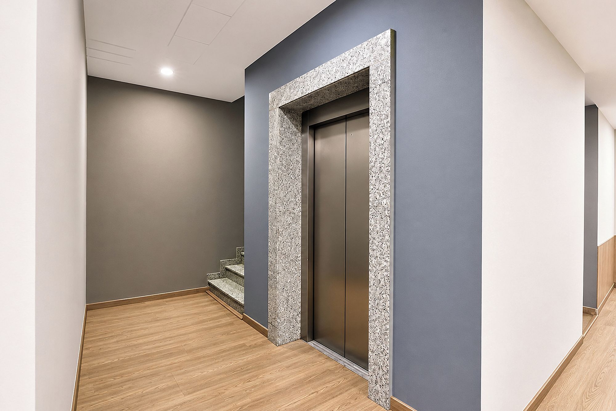 A hallway with a wooden floor, a grey wall, and a door with a textured frame.