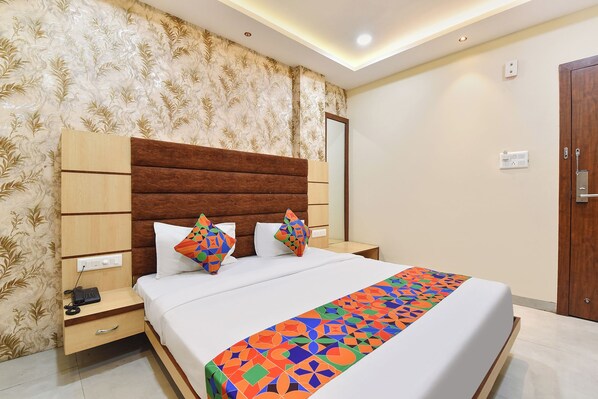 A hotel room with a bed, a bedside table with a phone, a nightstand, a wooden door, and a patterned wallpaper.