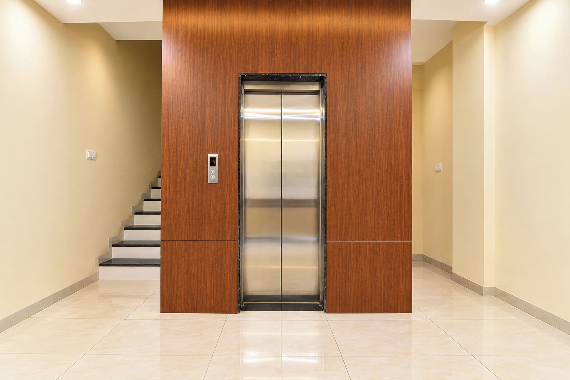 An elevator with a digital display, flanked by wooden panels, in a hallway with a tiled floor and a staircase to the left.