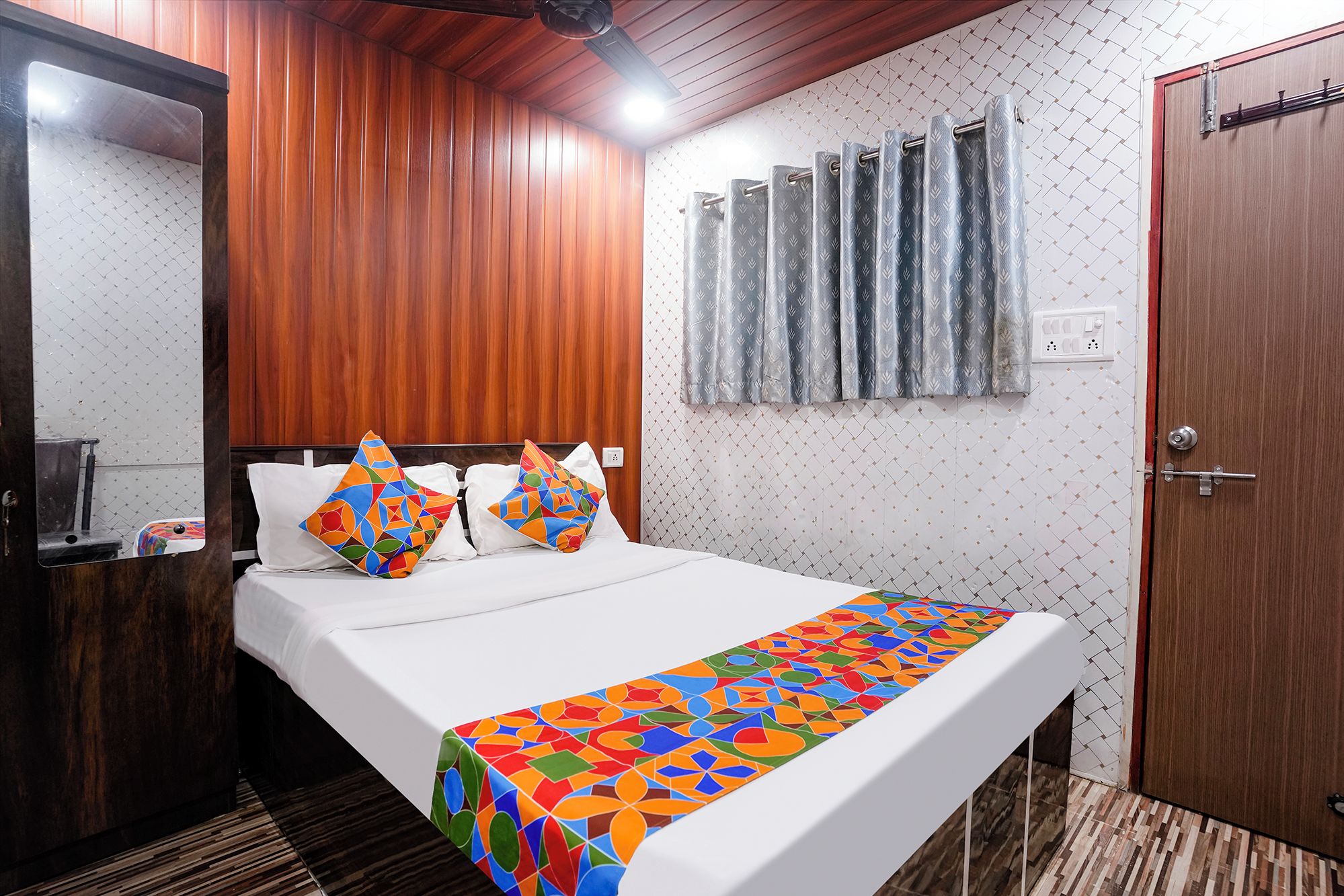 Ein Hotelzimmer mit Holzbalkendecke, einem Bett mit weißen Leinentüchern, bunten Kissen, einer gemusterten Tagesdecke und einem Fenster mit Vorhängen.