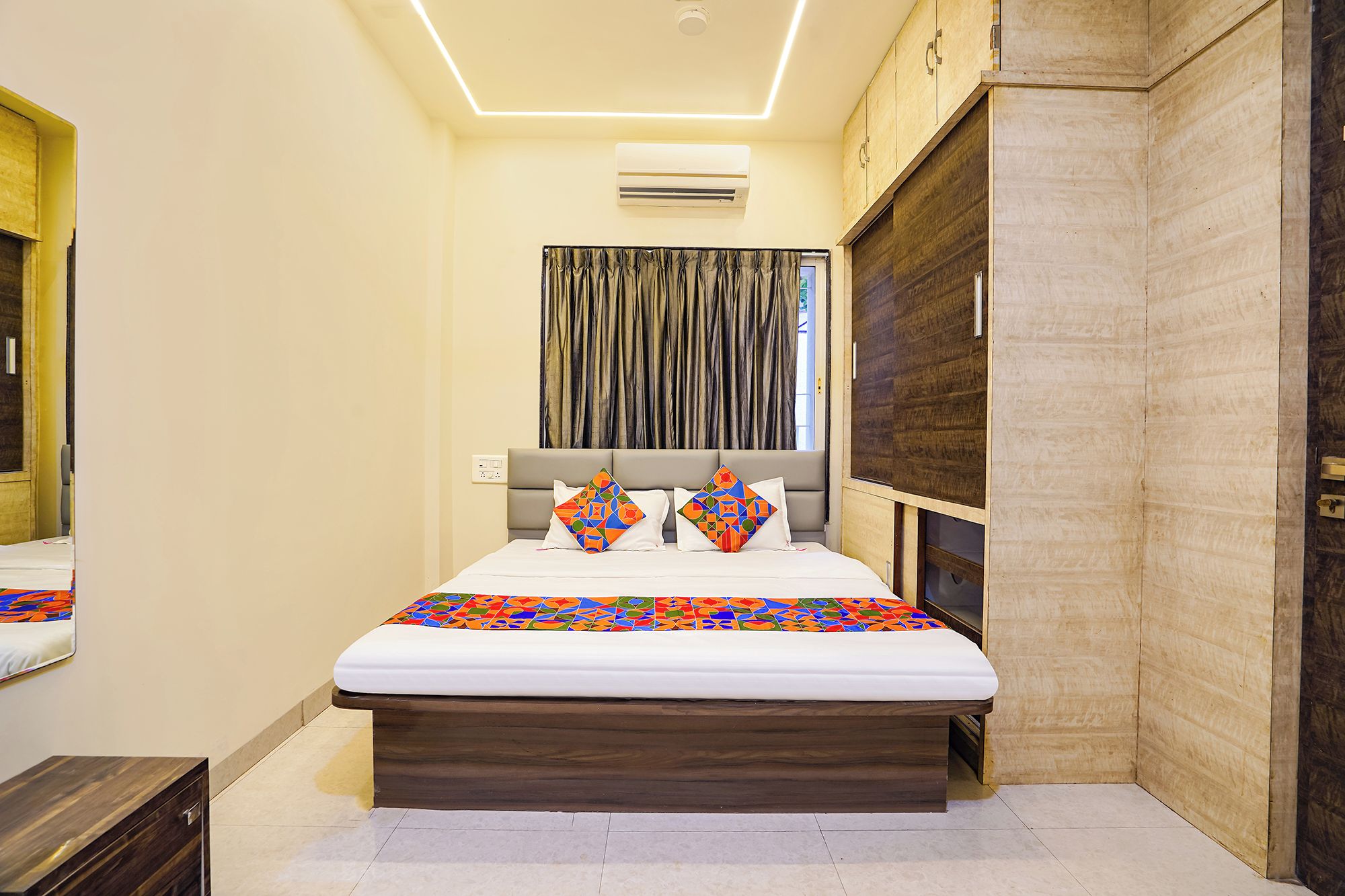 A bedroom with a bed, a headboard, a window with curtains, an air conditioning unit, and a wooden wardrobe.
