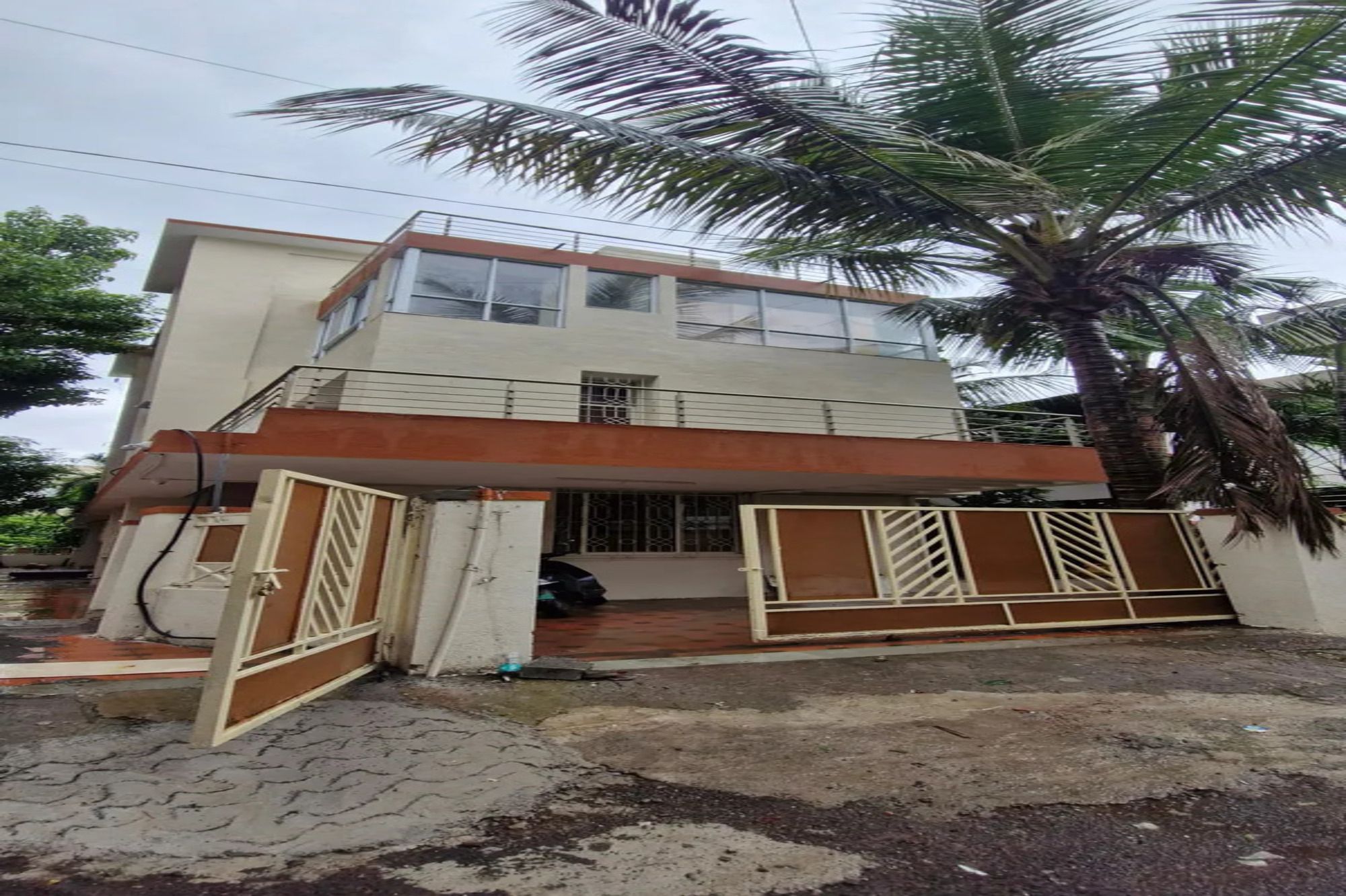 Un edificio de dos pisos con entrada protegida por un portón, un balcón visible y una palmera en primer plano.