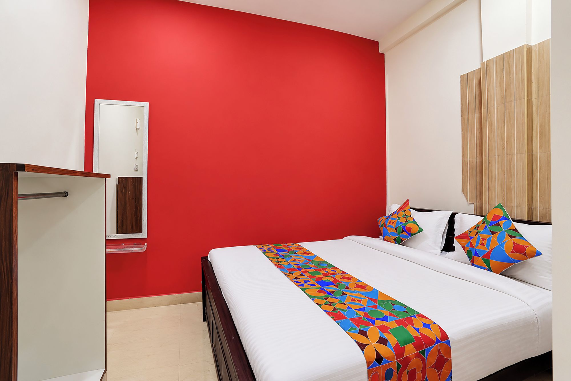 A hotel room with a red accent wall, a bed with a colorful geometric bedspread, and a wooden wardrobe.