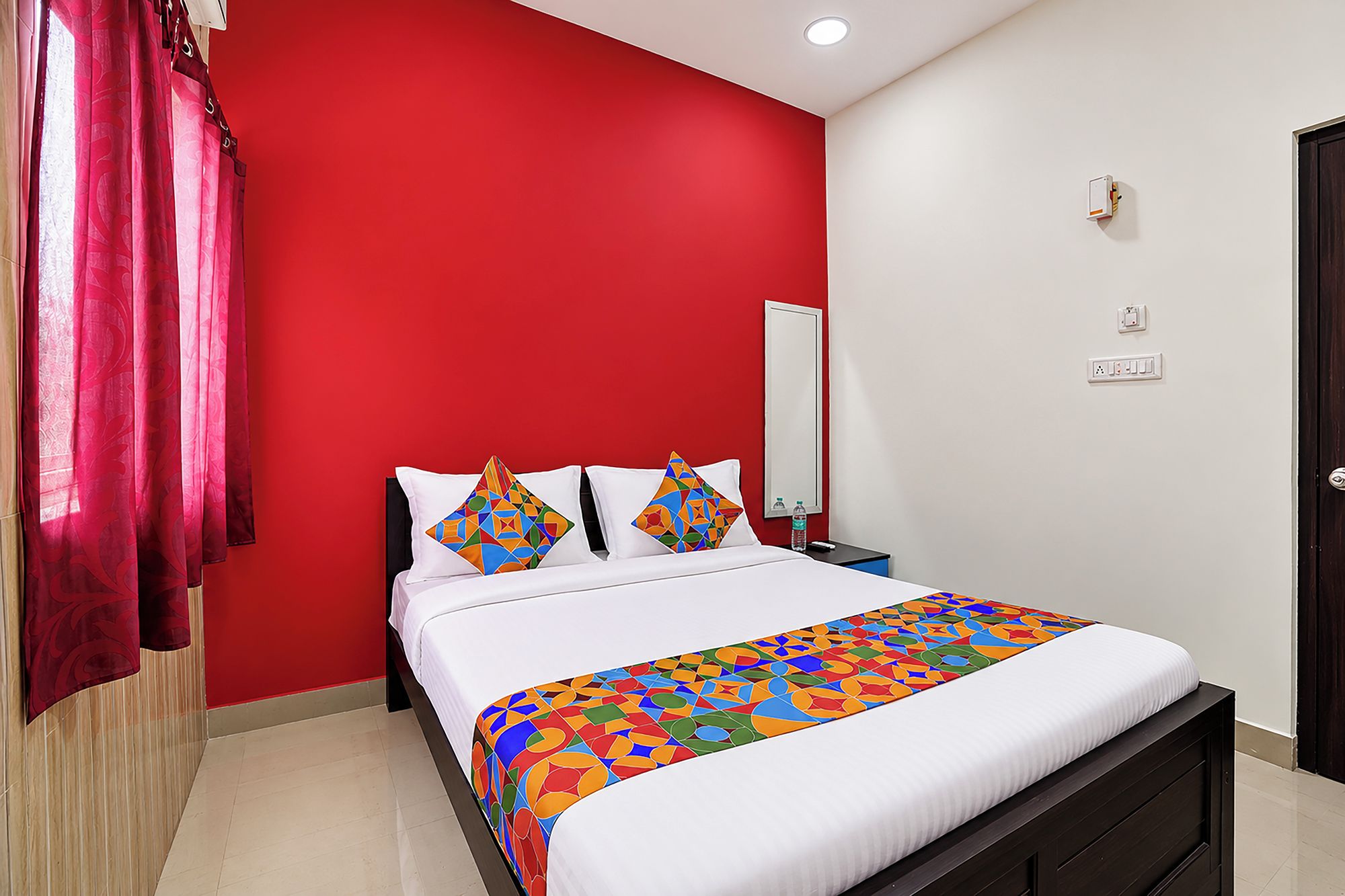 A hotel room with a red accent wall, a bed with white bedding, two colorful geometric pillows, a bedside table with a water bottle, and a door with a handle.