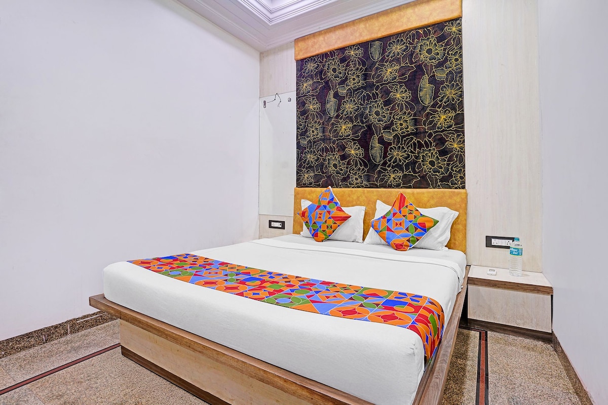 A hotel room with a bed, a headboard with a floral pattern, two pillows with geometric designs, and a bedside table with a water bottle.