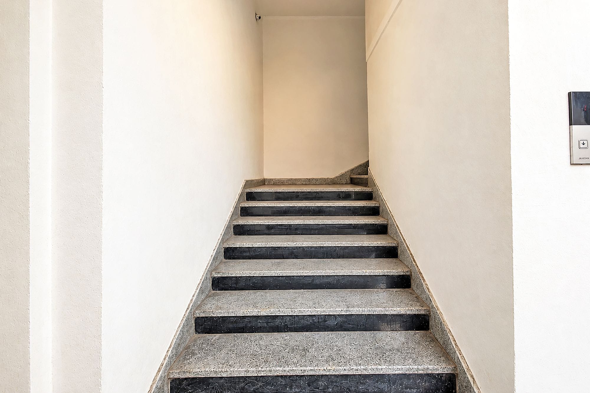 Eine Treppe mit schwarzen Stufen, die nach oben führt, weiße Wände und ein Lichtschalter an der rechten Wand.