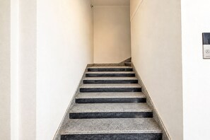 A staircase with black steps leading upwards, white walls, and a light switch on the right wall. - FabHotel Living Tree Suites (Hyderabad)