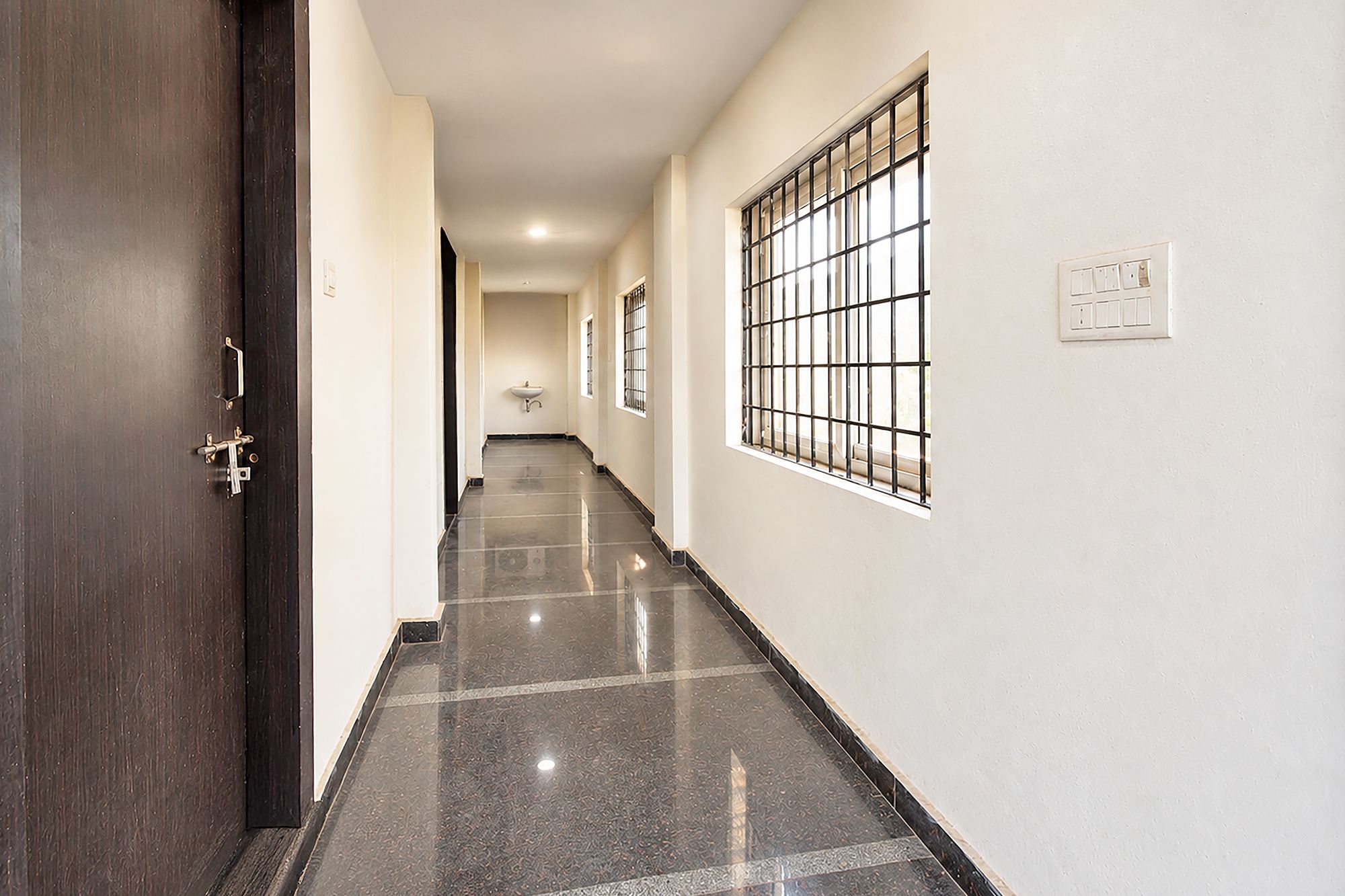 A corridor with a dark wooden door, a white wall, a window with metal bars, and a light switch.