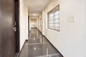 A corridor with a dark wooden door, a white wall, a window with metal bars, and a light switch. - FabHotel Living Tree Suites (Hyderabad)