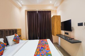 Ein Hotelzimmer mit Bett, Wandfernseher, Schreibtisch mit Flachbildfernseher und einem Vorhangfenster.