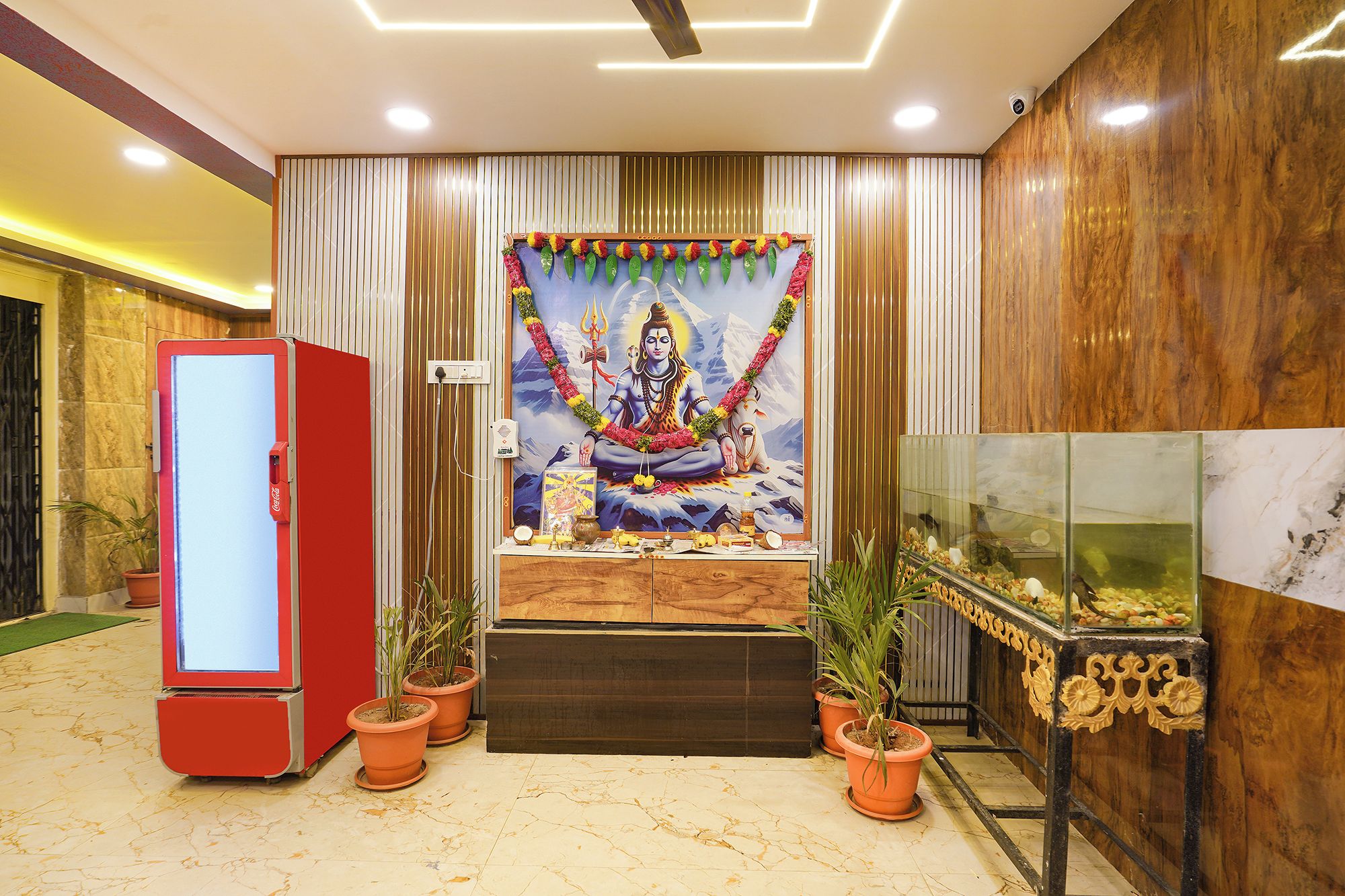 A room with a mural of a deity, a red refrigerator, potted plants, and a fish tank.