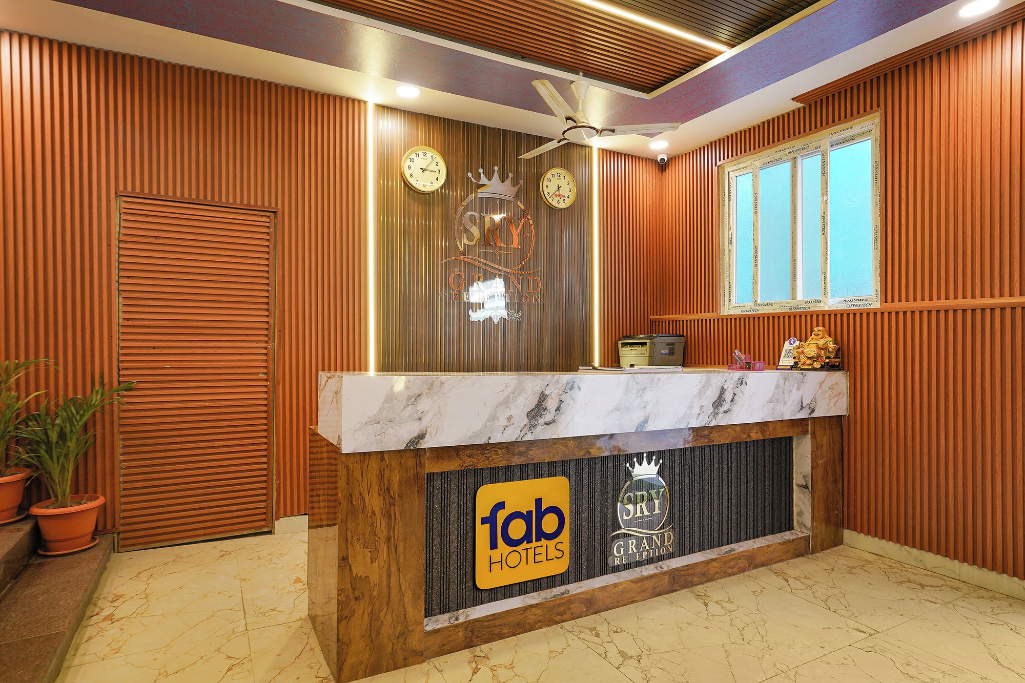 A hotel reception desk with a marble top, two clocks, a ceiling fan, and a window with blinds.