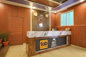 A hotel reception desk with a marble top, two clocks, a ceiling fan, and a window with blinds. - FabHotel Sry Grand (Hyderabad)