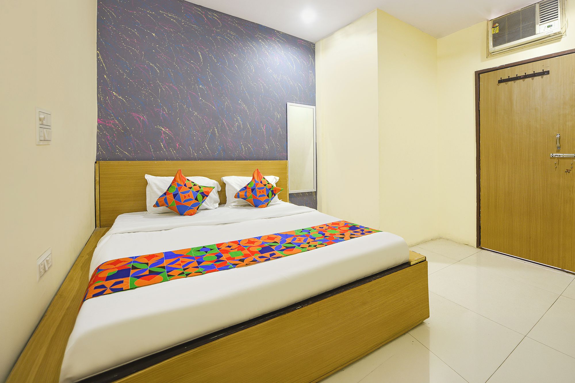 Habitación de hotel con una cama, cabecera de madera, colcha de diseño geométrico y pared lisa con textura, además de una puerta de madera.