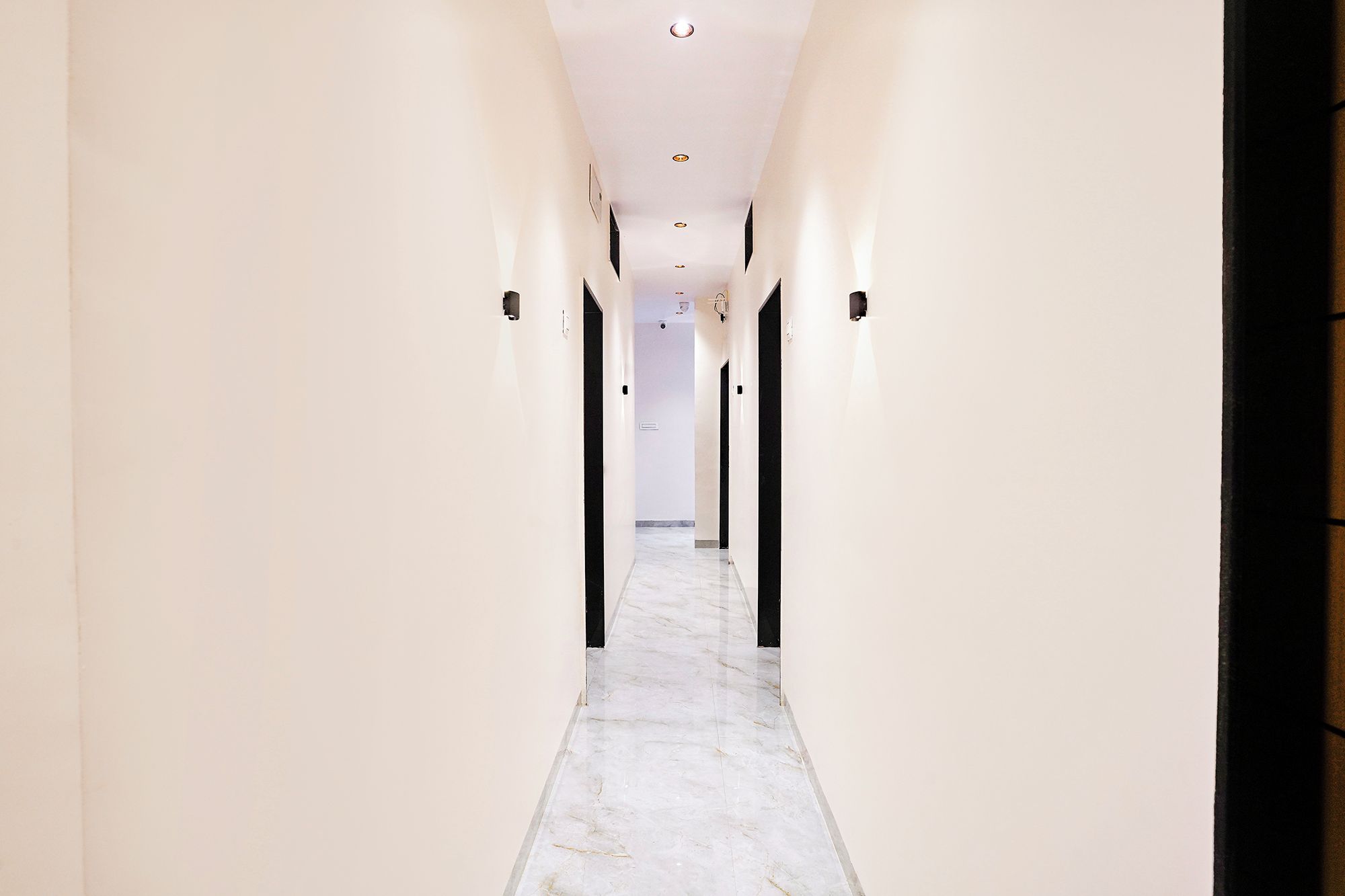 Un pasillo largo y estrecho con paredes blancas y suelo de mármol, que conduce a una habitación con una puerta oscura.