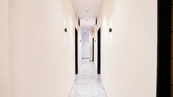 Un pasillo largo y estrecho con paredes blancas y suelo de mármol, que conduce a una habitación con una puerta oscura.