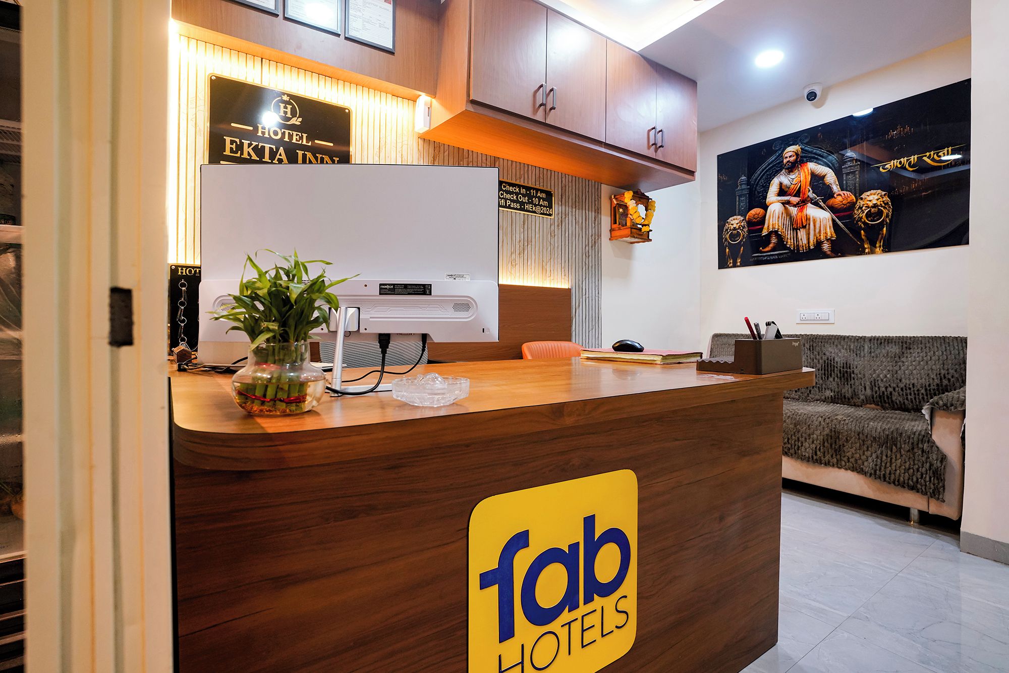 A hotel reception desk with a sign for 'fab hotels,' a plant, a computer, and a couch in the background.