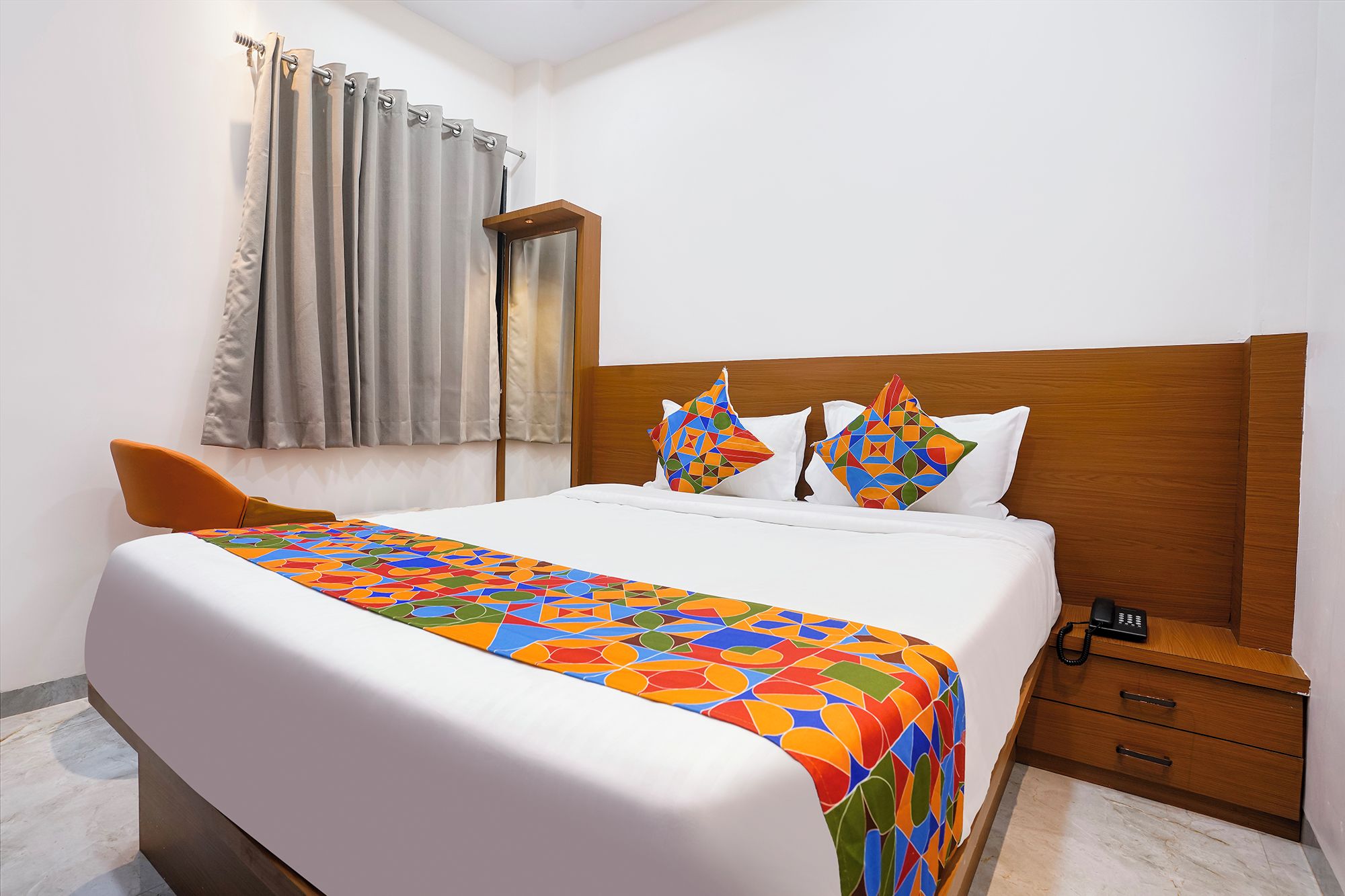 A hotel room with a bed, a bedside table with a phone, a chair, and a window with curtains.