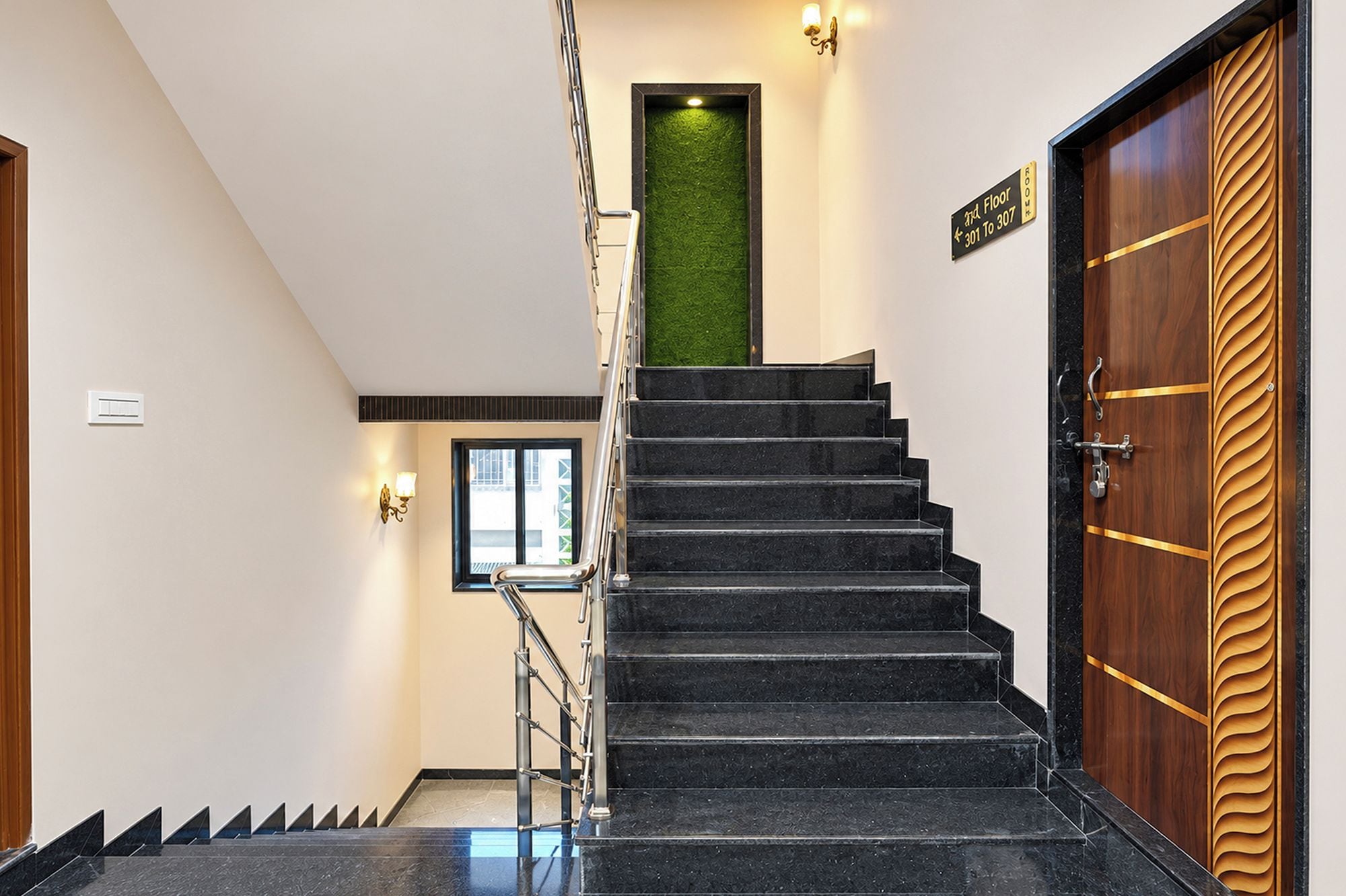 Una escalera con peldaños negros que conduce a una pared verde, una puerta de madera con una franja dorada y un cartel que indica los números de habitación 301 a 307.