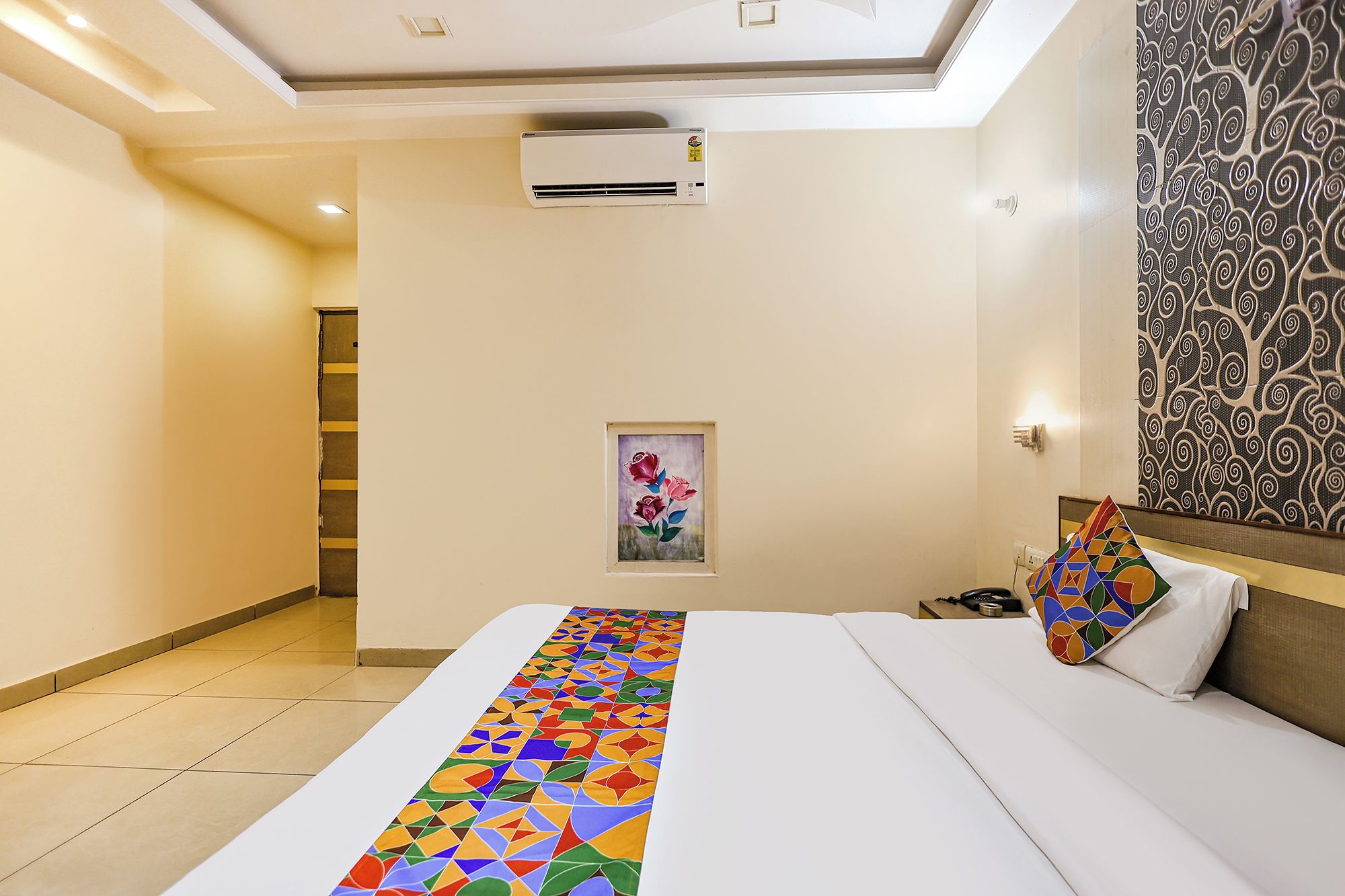 A hotel room with a bed, a painting, a patterned wall, and an air conditioning unit.