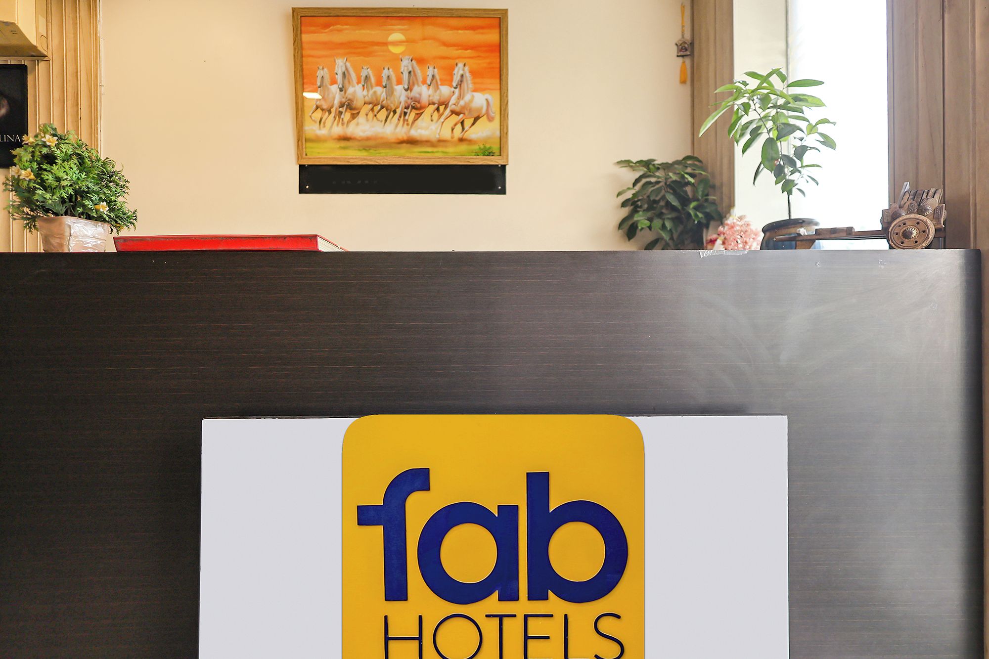 A reception desk with a sign for Fab Hotels, a painting of horses, and potted plants.