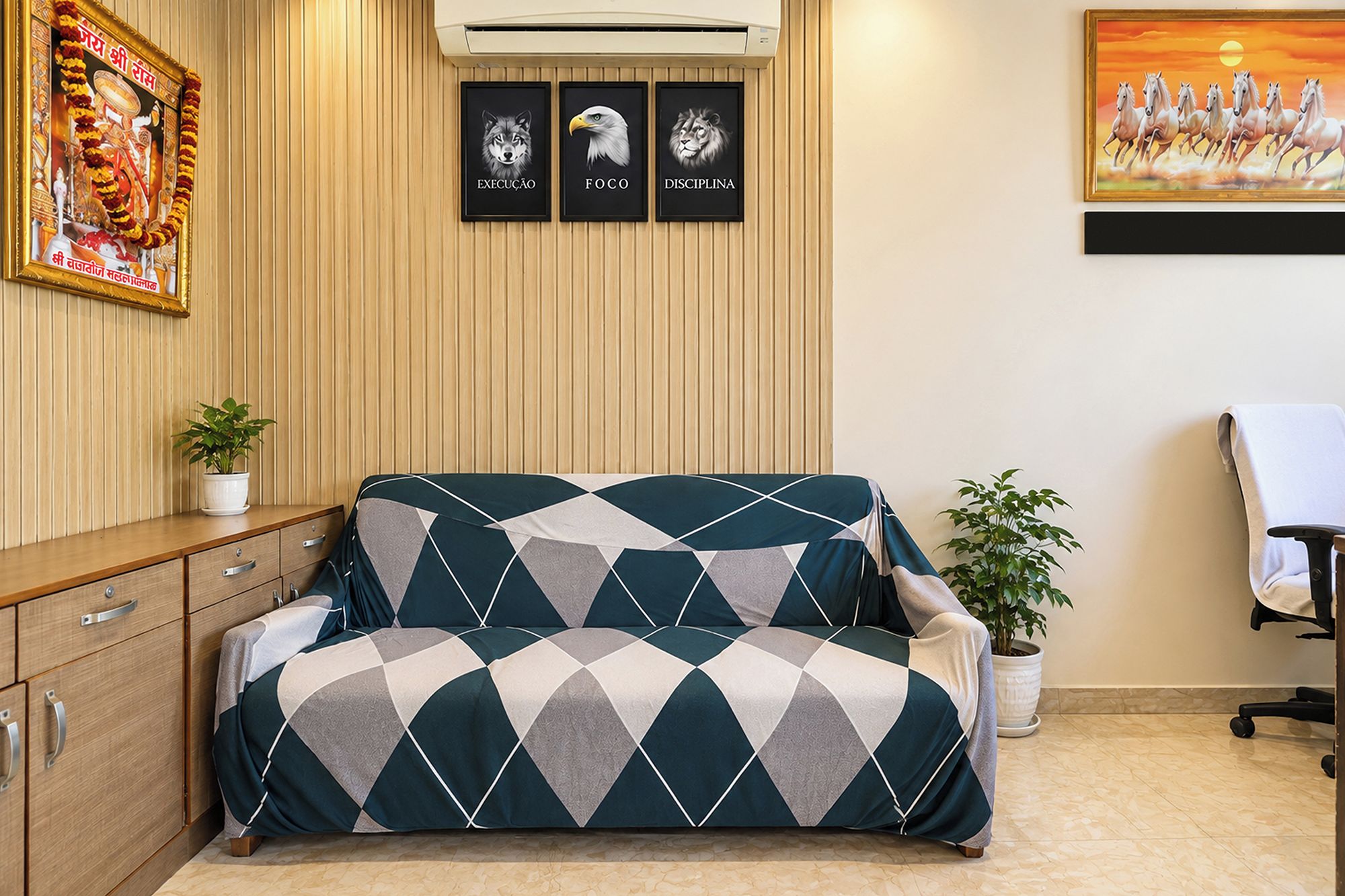 A room with a sofa, a desk, a chair, a potted plant, and framed artwork on the wall.