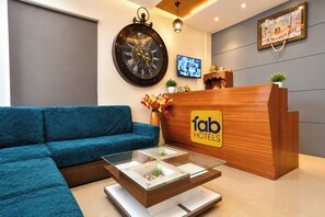 Un espace d’accueil d’hôtel avec un canapé bleu, un comptoir d’accueil en bois, une horloge murale et un écran de télévision.