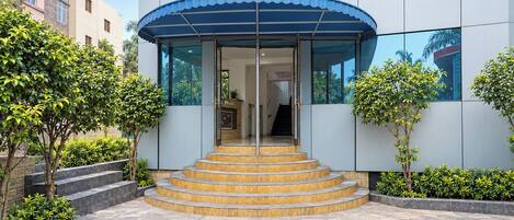 A modern building entrance with a curved blue awning, a set of stairs leading to the entrance, and surrounding greenery.