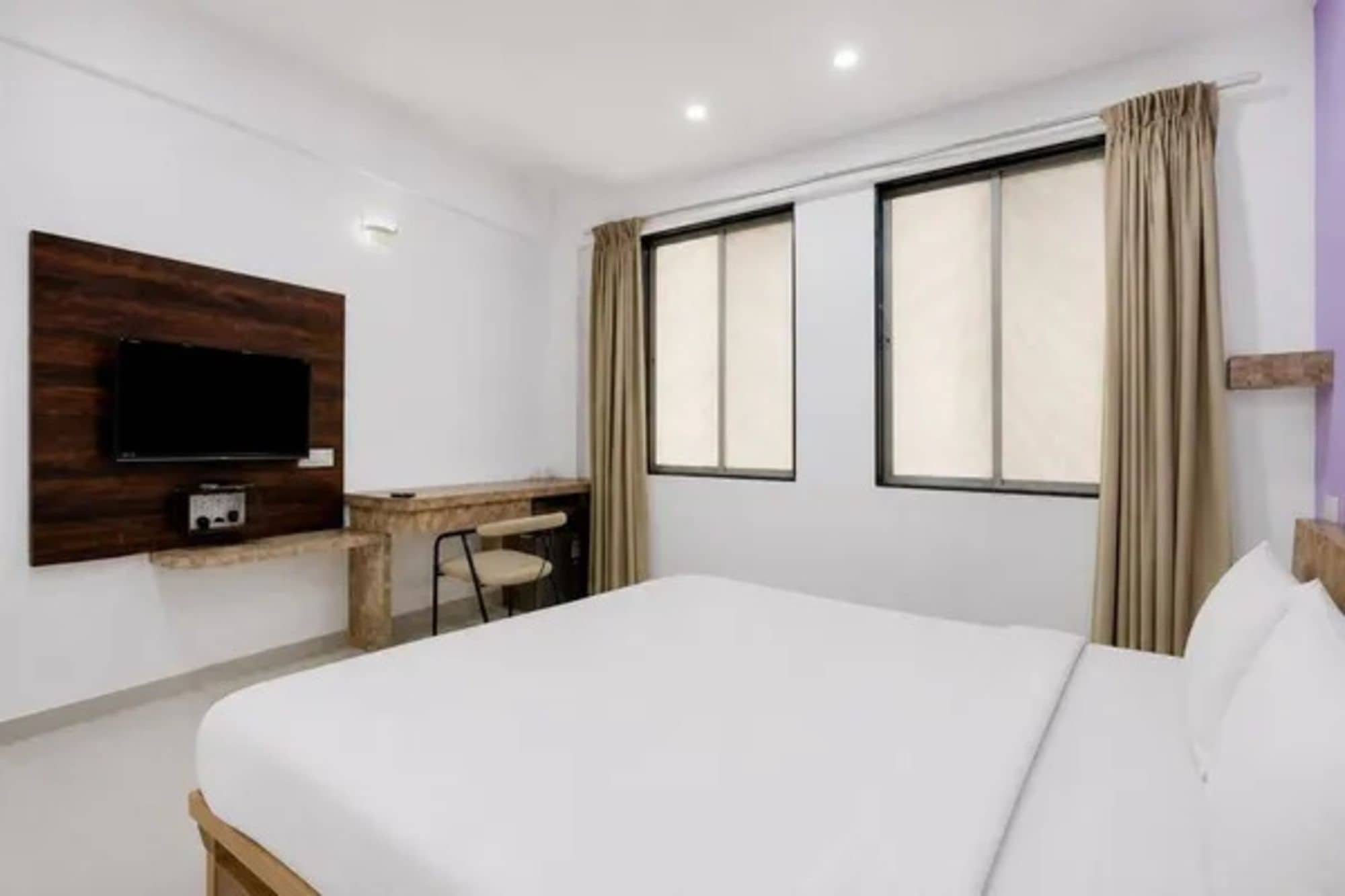 Ein Hotelzimmer mit einem Bett, einem Schreibtisch, einem Stuhl, einem Fernseher und zwei Fenstern mit Vorhängen.