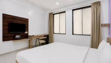 A hotel room with a bed, a desk, a chair, a television, and two windows with curtains.