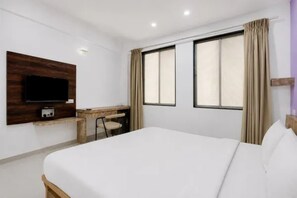Habitación de hotel con cama, escritorio, silla, televisor y dos ventanas con cortinas.